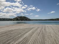 there is a wooden floor by the water on top of it with the beach and hills in the background