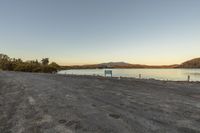 an empty lot on the side of a lake at dusk by a bench and mountain range