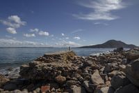 a man is standing near the shore line by some rocks and water with a mountain in the background