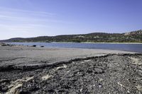 a rocky shore along a lake with mountains in the distance, with dirt and rocks strewn on the beach