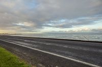 a street by the sea with waves on the water next to it and a long grass field beside a road that has a large patch of land under the road