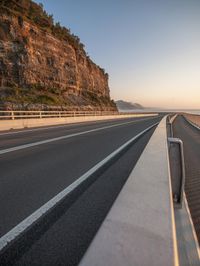 a highway near some big rocks and grass and sky at sunset behind it with the ocean