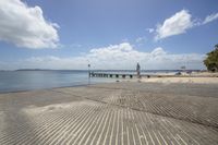 a walkway along the water near a beach area with benches and a pier with a sky background