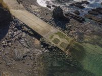 a walkway that leads to the water between the beach and rocks near a shoreline with a rock lighthouse behind it