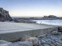 a skateboarder stands on the edge of a stone ramp overlooking the ocean at twilight