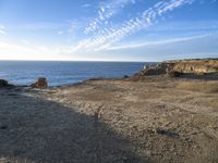 two people walking toward the ocean near a rocky shore, in front of some clouds