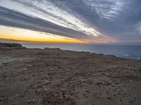 a bench sitting on the shore of a beach near the ocean at sunset during an overcast sky