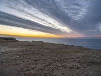 a bench sitting on the shore of a beach near the ocean at sunset during an overcast sky