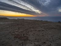 a bench sitting on the shore of a beach near the ocean at sunset during an overcast sky