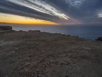 a bench sitting on the shore of a beach near the ocean at sunset during an overcast sky