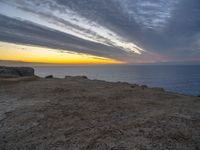 a bench sitting on the shore of a beach near the ocean at sunset during an overcast sky