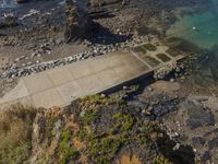 the aerial view of an empty concrete area near the ocean, on a rocky beach