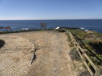 a dirt road with a sign next to the ocean by some wooden fences near a cliff