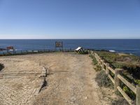 a dirt road with a sign next to the ocean by some wooden fences near a cliff