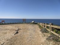 a dirt road with a sign next to the ocean by some wooden fences near a cliff