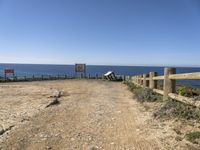 a dirt road with a sign next to the ocean by some wooden fences near a cliff