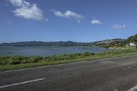 a street near a body of water near some houses on a hill side line with hills and a field in front