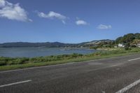 a street near a body of water near some houses on a hill side line with hills and a field in front