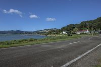 a street near a body of water near some houses on a hill side line with hills and a field in front