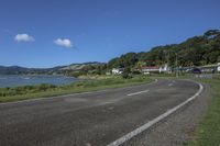 a street near a body of water near some houses on a hill side line with hills and a field in front