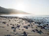 large number of small rocks strewn on the beach near water and mountains behind them with sun shining down in the sky