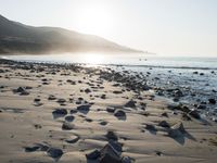 large number of small rocks strewn on the beach near water and mountains behind them with sun shining down in the sky