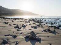 large number of small rocks strewn on the beach near water and mountains behind them with sun shining down in the sky