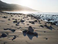 large number of small rocks strewn on the beach near water and mountains behind them with sun shining down in the sky