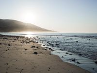 large number of small rocks strewn on the beach near water and mountains behind them with sun shining down in the sky