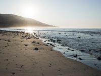 large number of small rocks strewn on the beach near water and mountains behind them with sun shining down in the sky