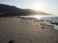 large number of small rocks strewn on the beach near water and mountains behind them with sun shining down in the sky