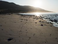 large number of small rocks strewn on the beach near water and mountains behind them with sun shining down in the sky