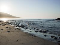 large number of small rocks strewn on the beach near water and mountains behind them with sun shining down in the sky