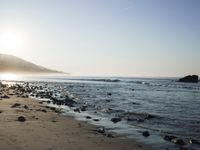 large number of small rocks strewn on the beach near water and mountains behind them with sun shining down in the sky