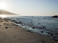 large number of small rocks strewn on the beach near water and mountains behind them with sun shining down in the sky