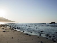 large number of small rocks strewn on the beach near water and mountains behind them with sun shining down in the sky