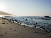 large number of small rocks strewn on the beach near water and mountains behind them with sun shining down in the sky