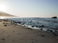 large number of small rocks strewn on the beach near water and mountains behind them with sun shining down in the sky