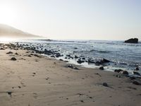 large number of small rocks strewn on the beach near water and mountains behind them with sun shining down in the sky