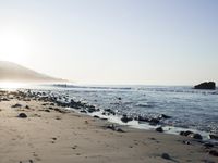 large number of small rocks strewn on the beach near water and mountains behind them with sun shining down in the sky