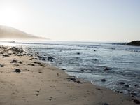 large number of small rocks strewn on the beach near water and mountains behind them with sun shining down in the sky