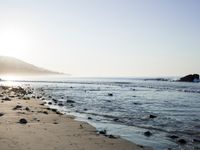 large number of small rocks strewn on the beach near water and mountains behind them with sun shining down in the sky