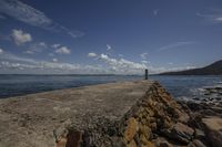 the water and rocks are beside the beach with clouds in the sky and a large rock wall