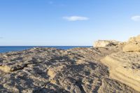 a rocky cliff near the ocean with some waves on it, and blue skies above