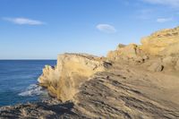 a rocky cliff near the ocean with some waves on it, and blue skies above