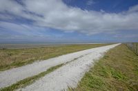 a gravel road next to an empty field with the ocean in the background and blue sky above