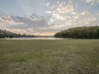 a field with some grass trees and water near by at sunrise sky and clouds and sun