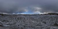 waves in ocean water under dark clouds and a hillside with mountains in background that appear to be covered by grey, stormy skies