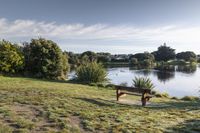 a wooden bench sitting on top of a grass covered field next to a lake and bushes