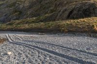 a lone sheep stands in the middle of a gravel field near the water and mountains